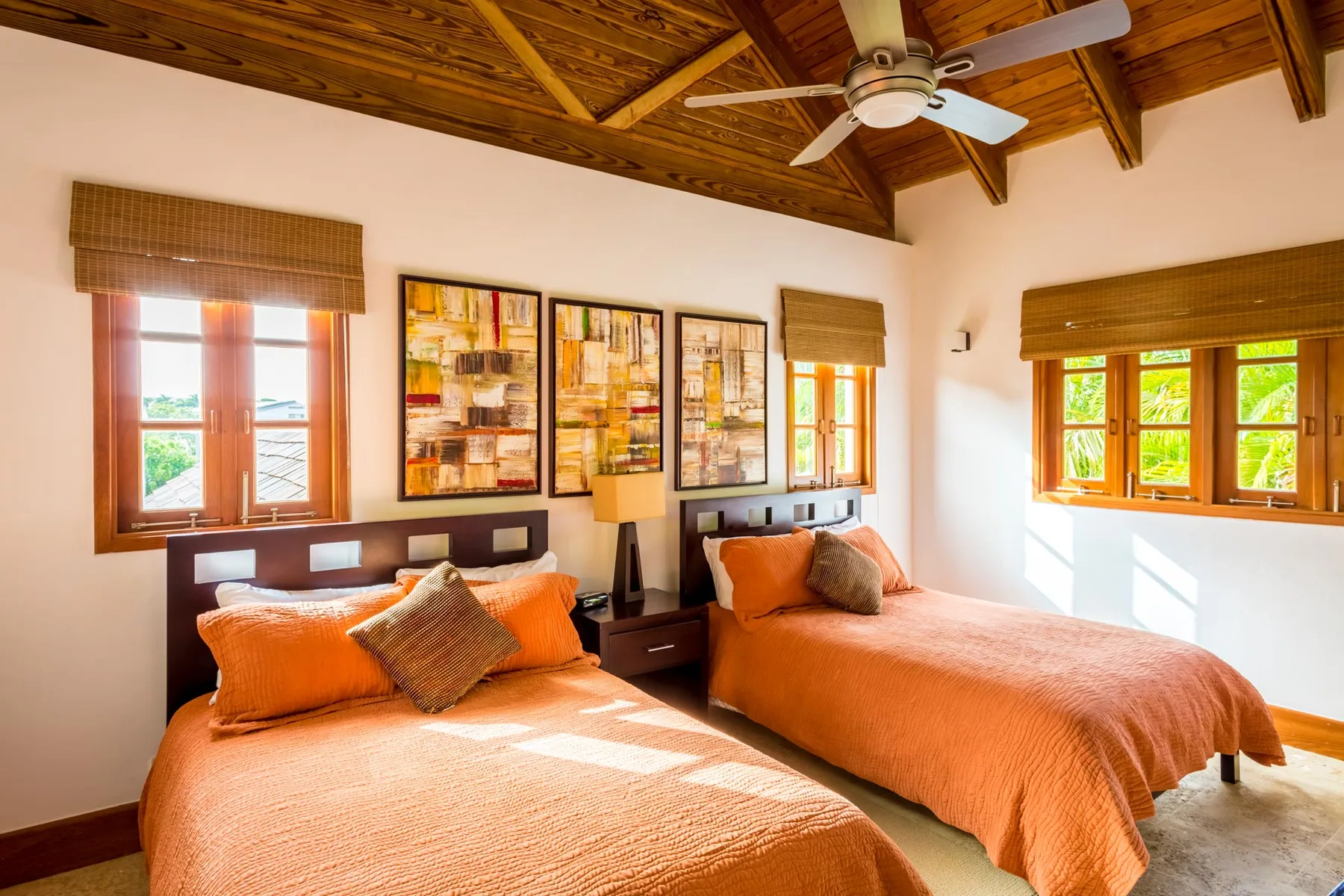 A bright bedroom with two beds, framed artwork, and windows beneath a wooden ceiling.