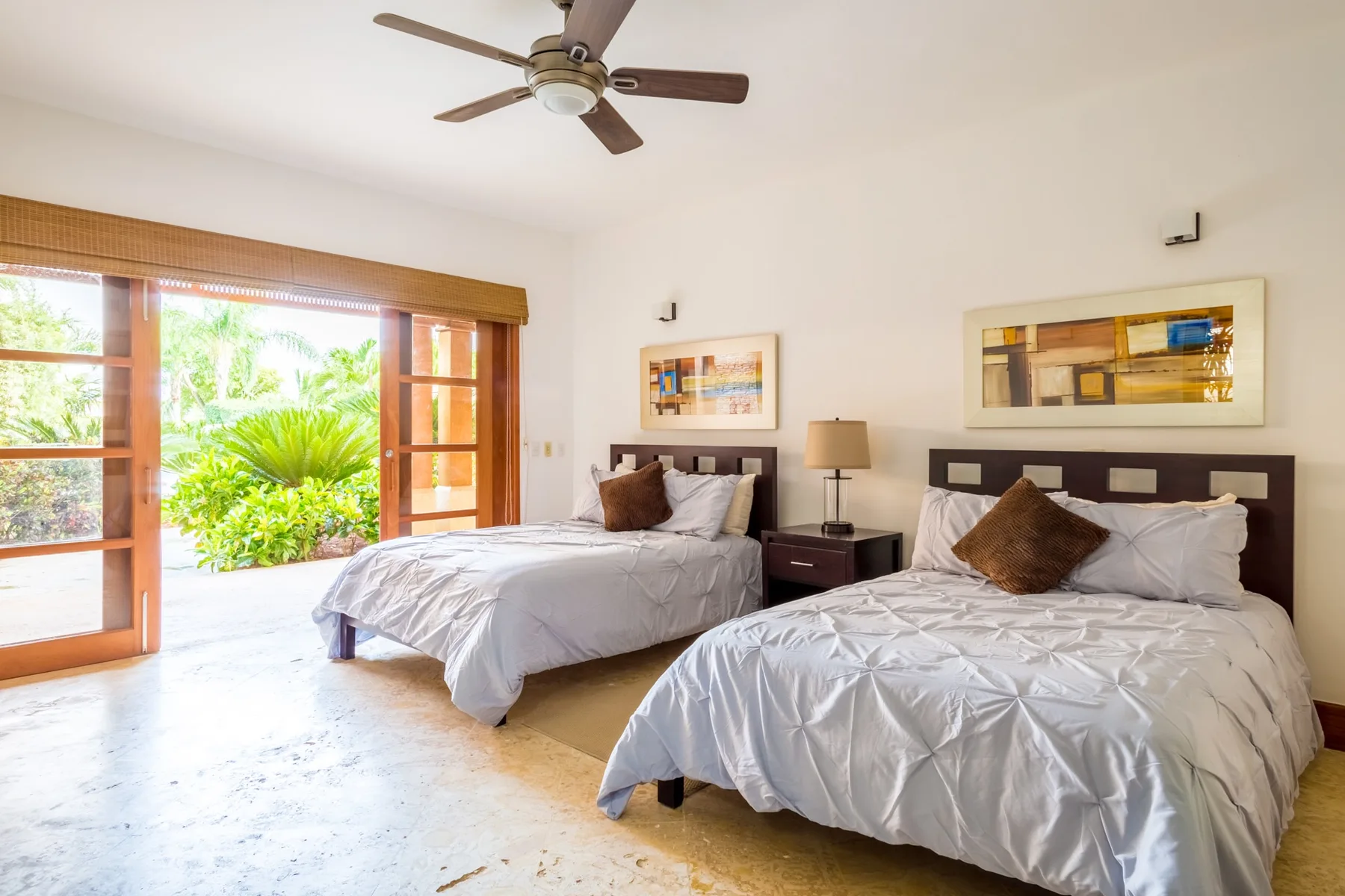 A bedroom with two beds, bedside tables, and an open view to greenery.