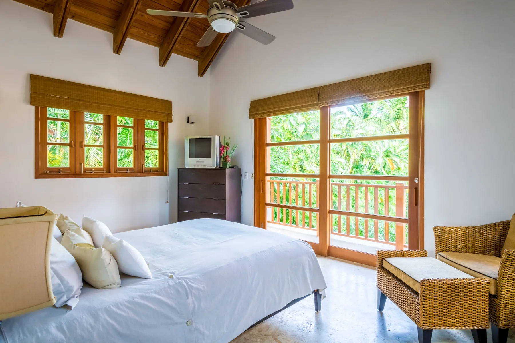 A bright bedroom with a large bed beside windows overlooking leafy greenery.