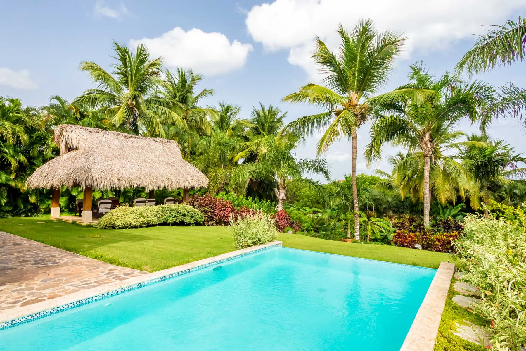 A swimming pool beside a thatched pavilion, surrounded by palms and tropical greenery.