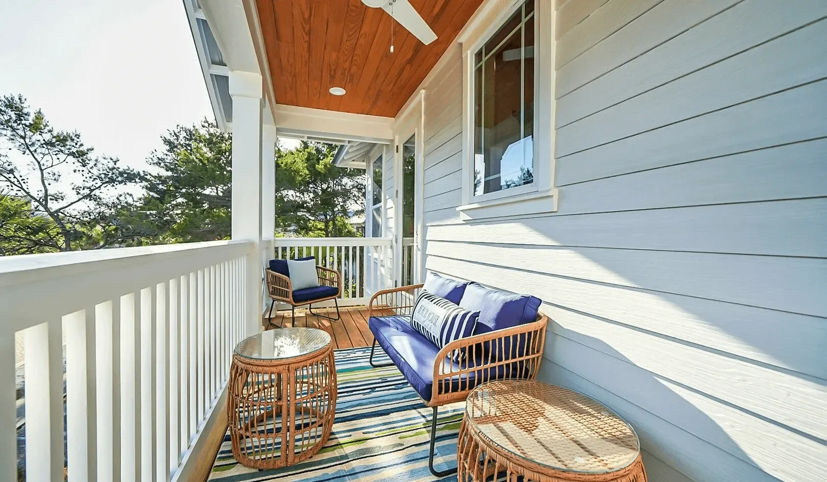 A covered porch with blue seating and a striped rug overlooks leafy trees.