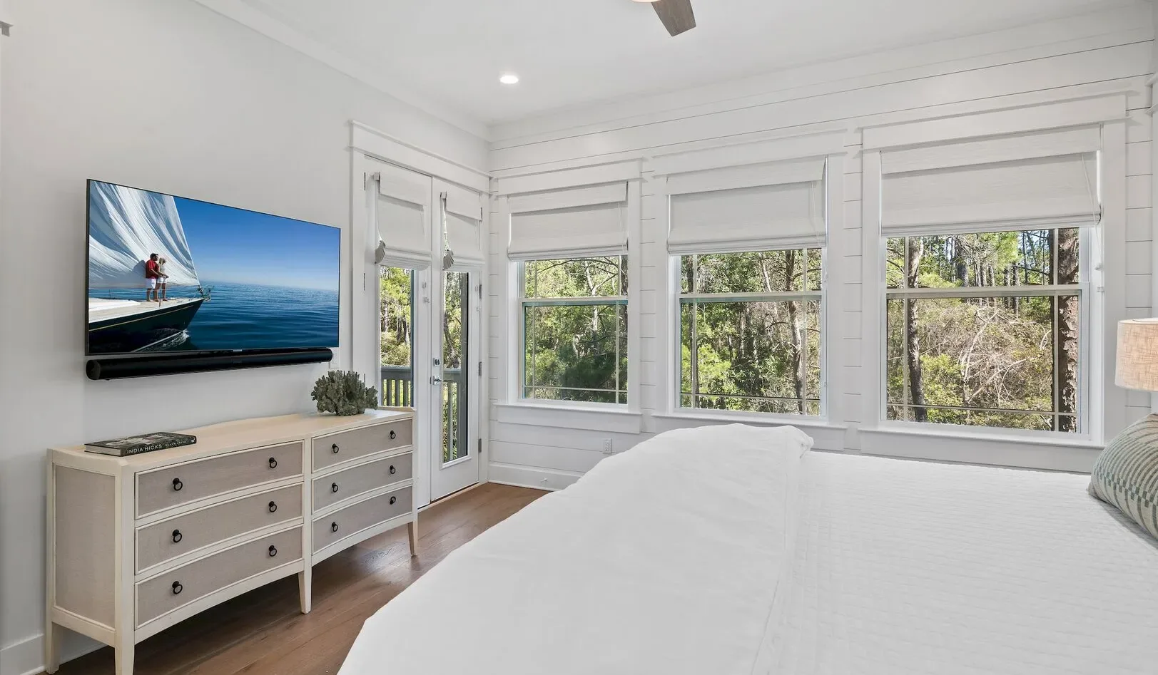 A bright bedroom with a bed, dresser, and large windows overlooking trees.