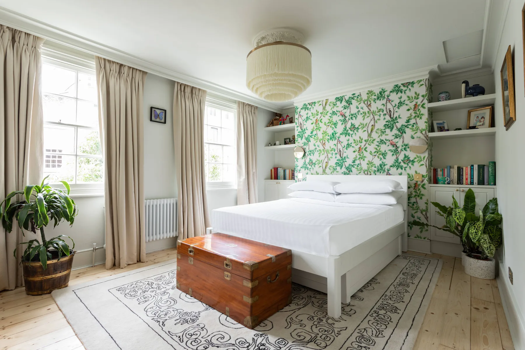 A bright bedroom with a white bed, patterned wallpaper, and tall windows.