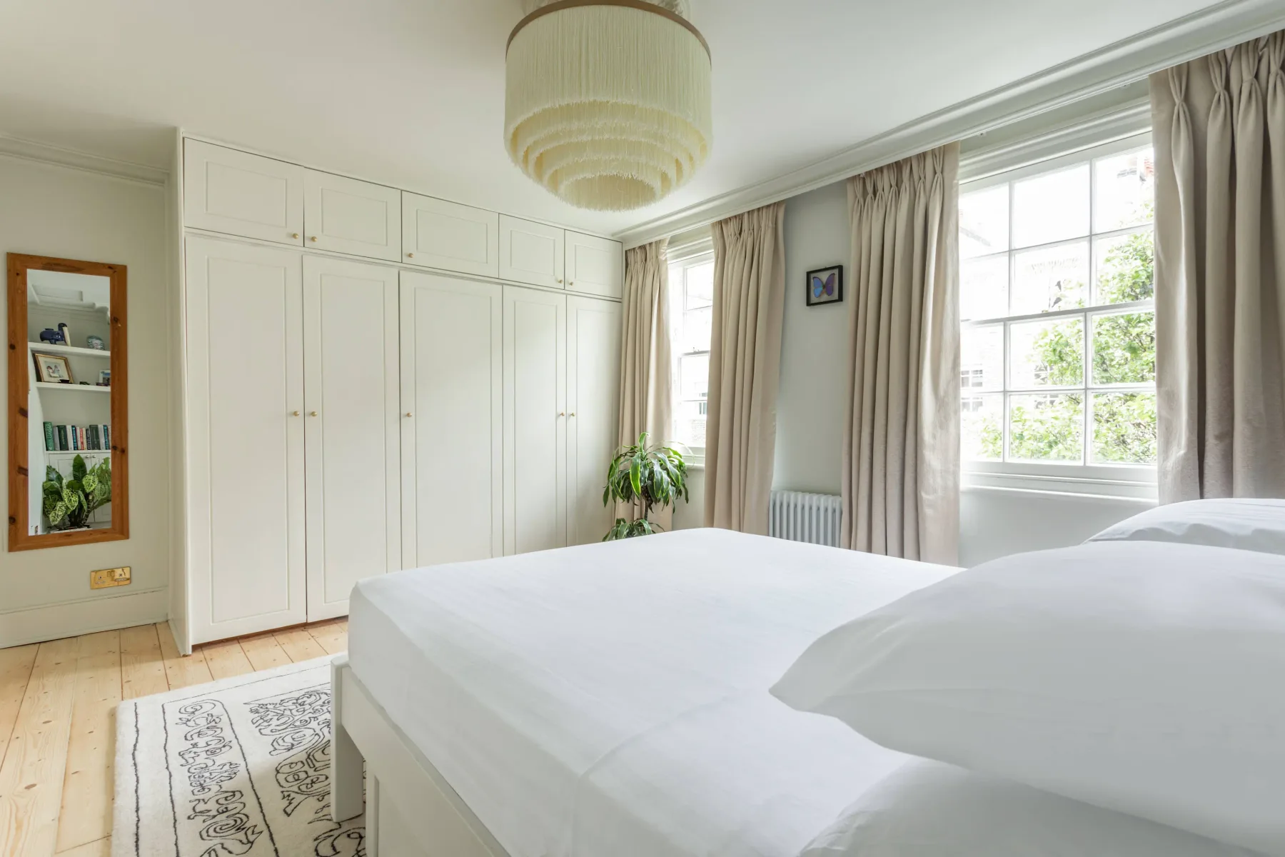 A bright bedroom with white bedding, built-in cabinets, and large curtained windows.