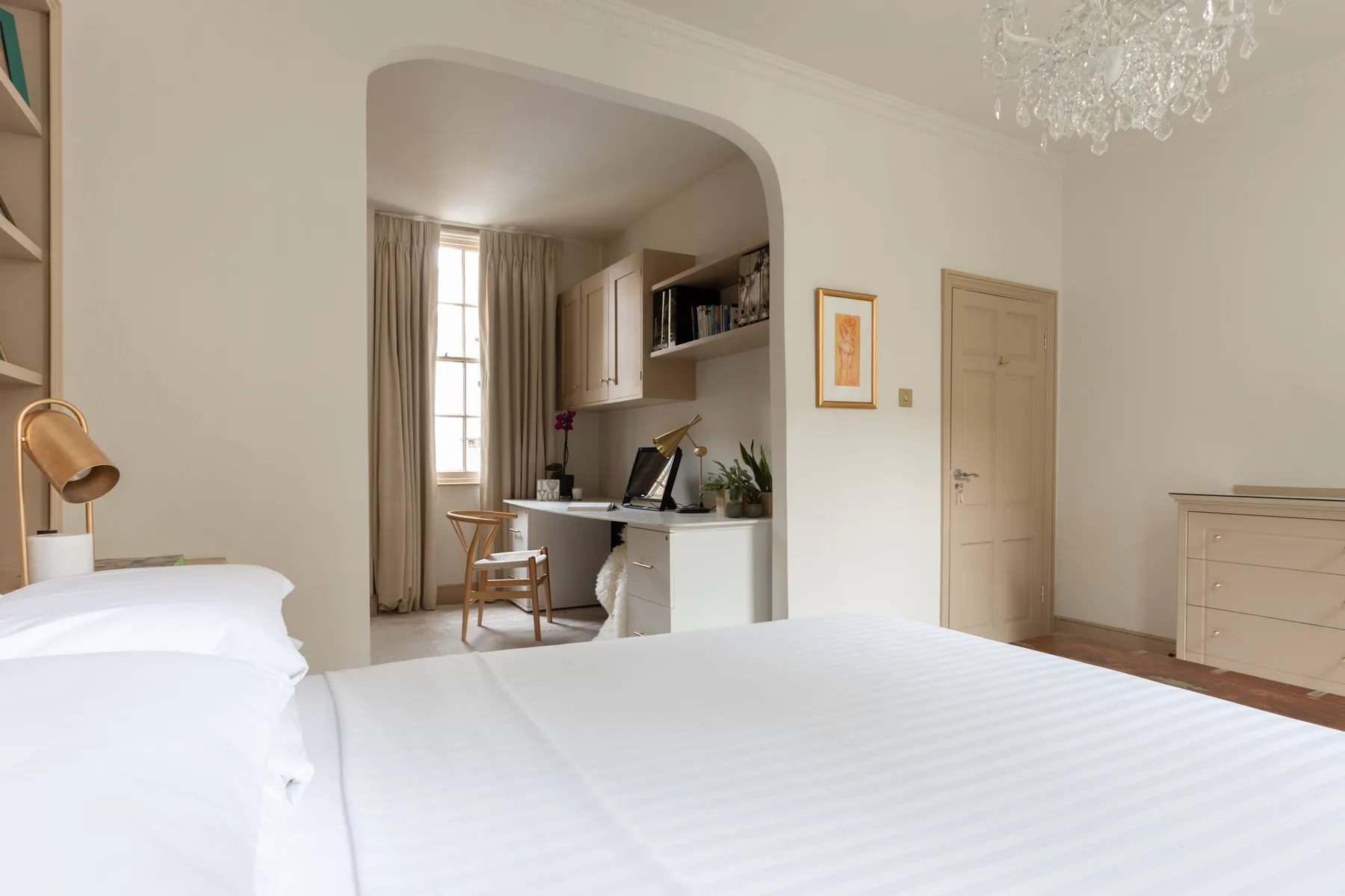 A bright bedroom with a white bed and a small kitchen area beyond an archway.