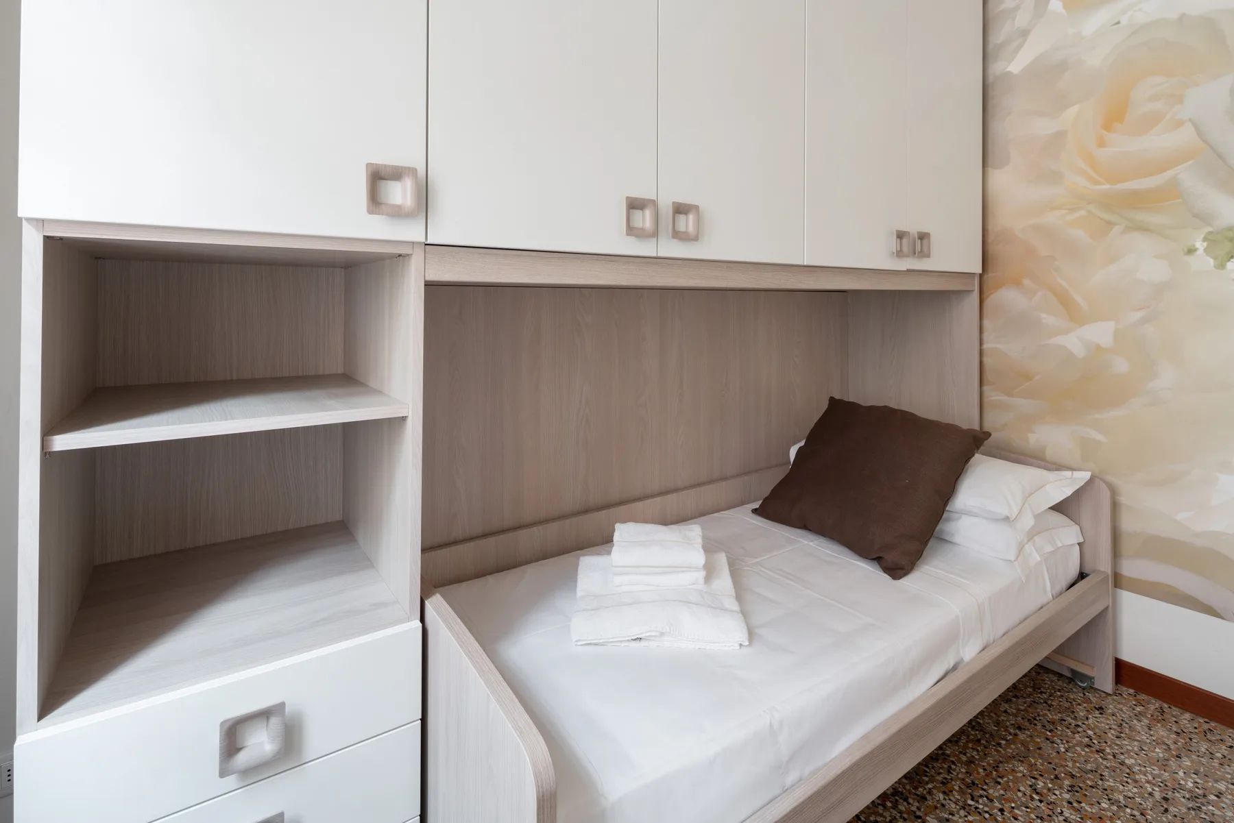 Una cama individual está bajo armarios empotrados, con un cojín marrón y una pared estampada.
