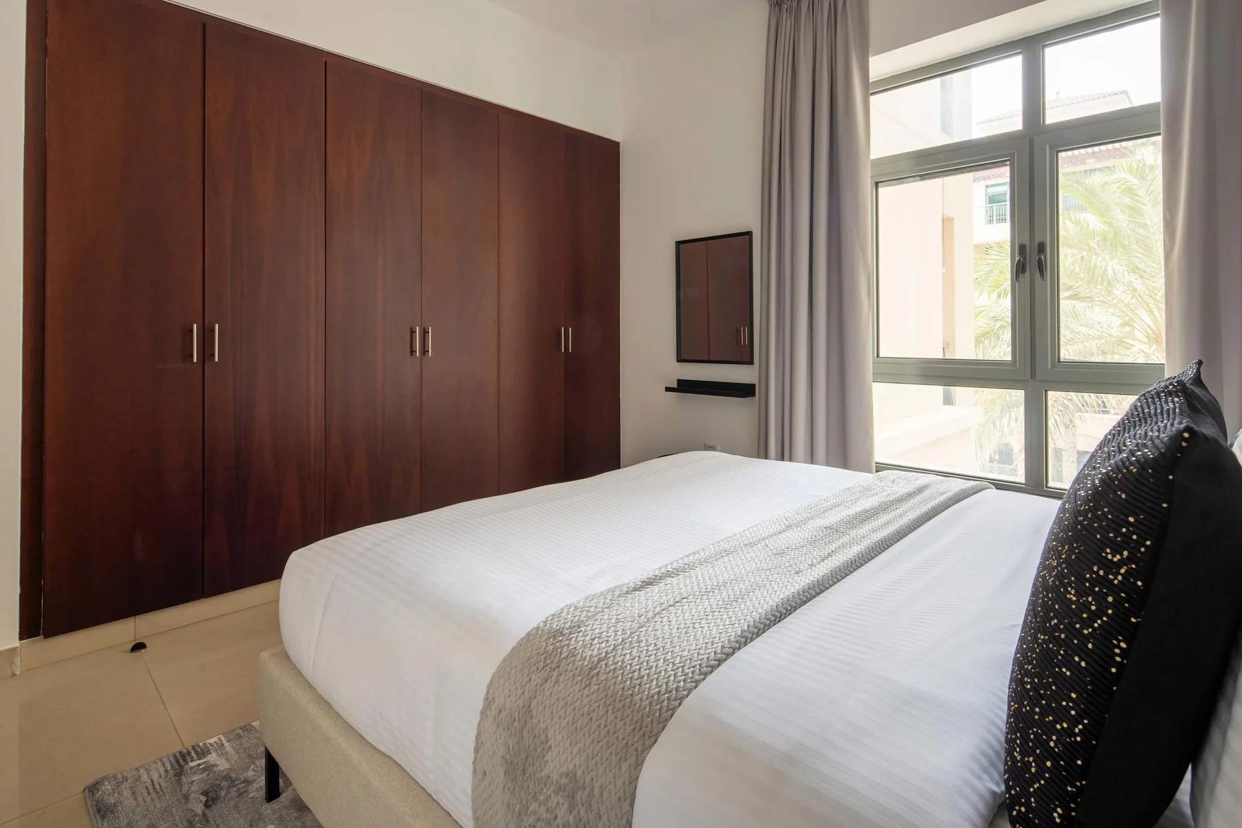 A bedroom with a white bed beside a large window and dark wood wardrobes.