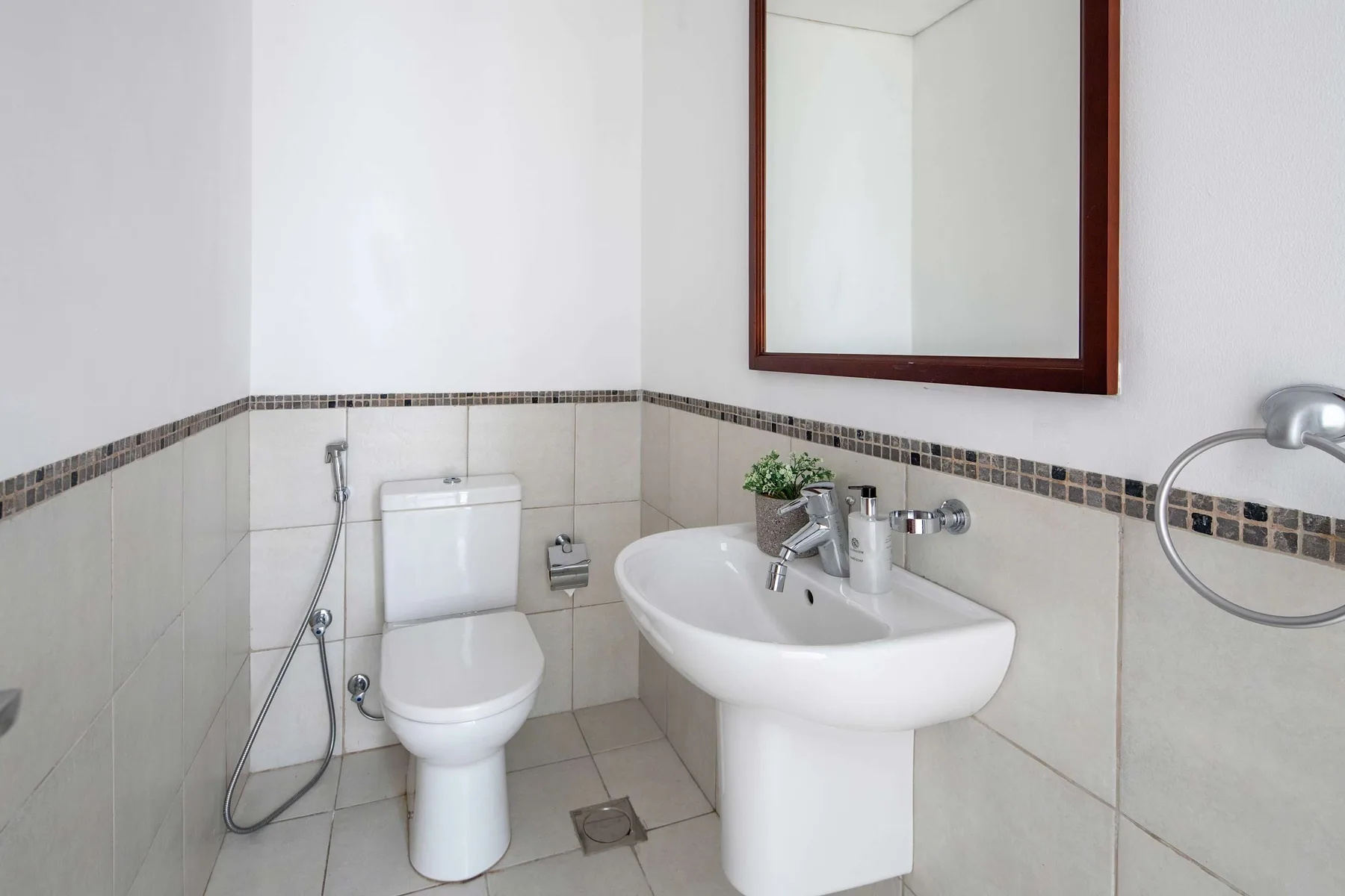 A tiled bathroom with a toilet, bidet sprayer, and pedestal sink beneath a mirror.