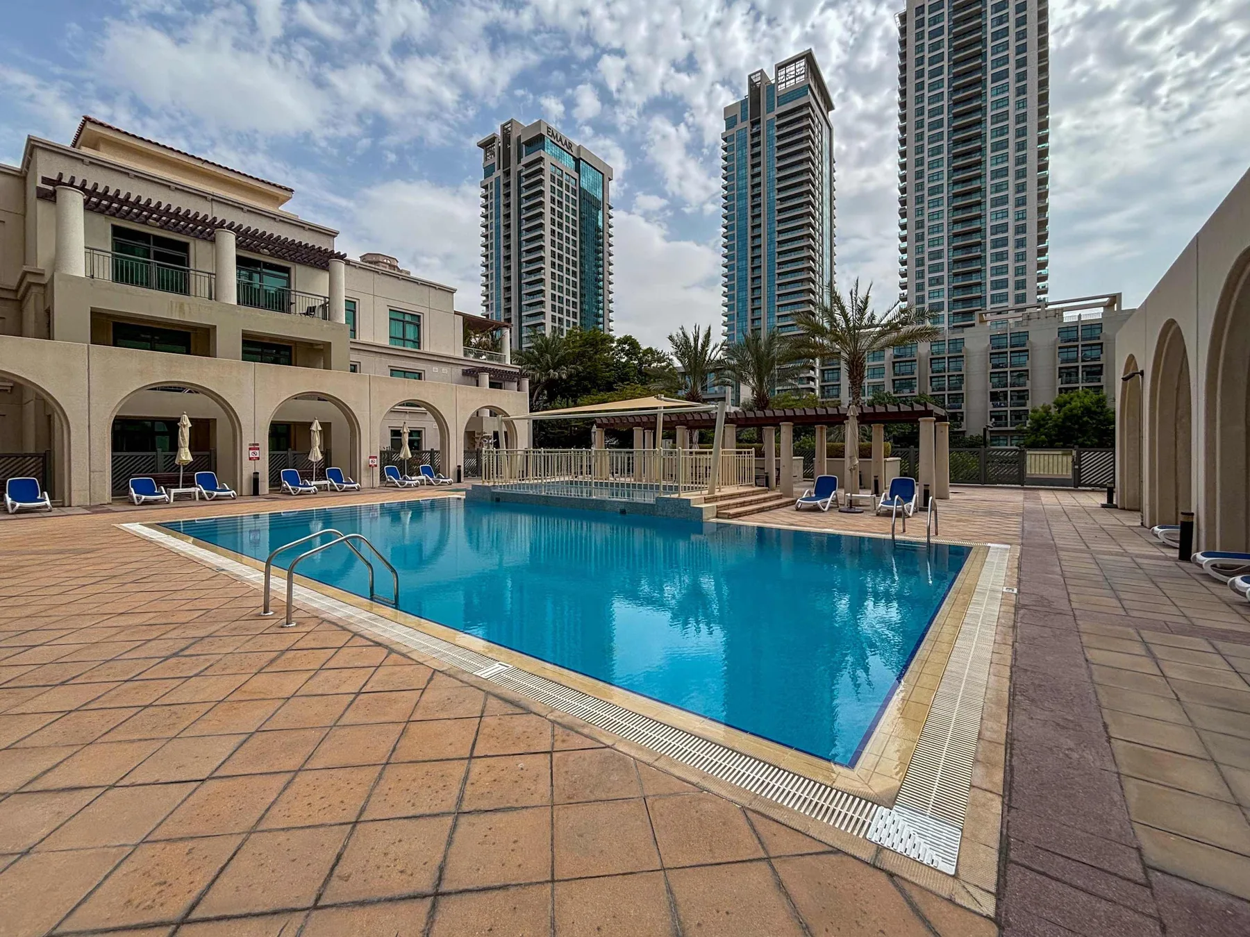An outdoor swimming pool sits among apartment towers, with lounge seating around the deck.