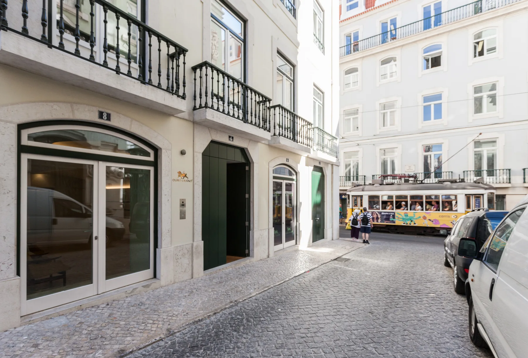 Una calle empedrada pasa entre edificios de tonos claros, con vehículos estacionados y un tranvía.