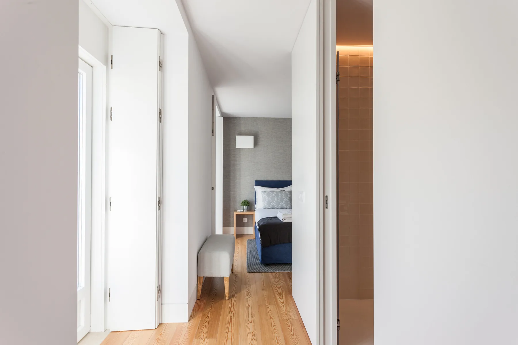 Se ve un dormitorio luminoso a través de una puerta abierta, con una cama y suelo de madera.