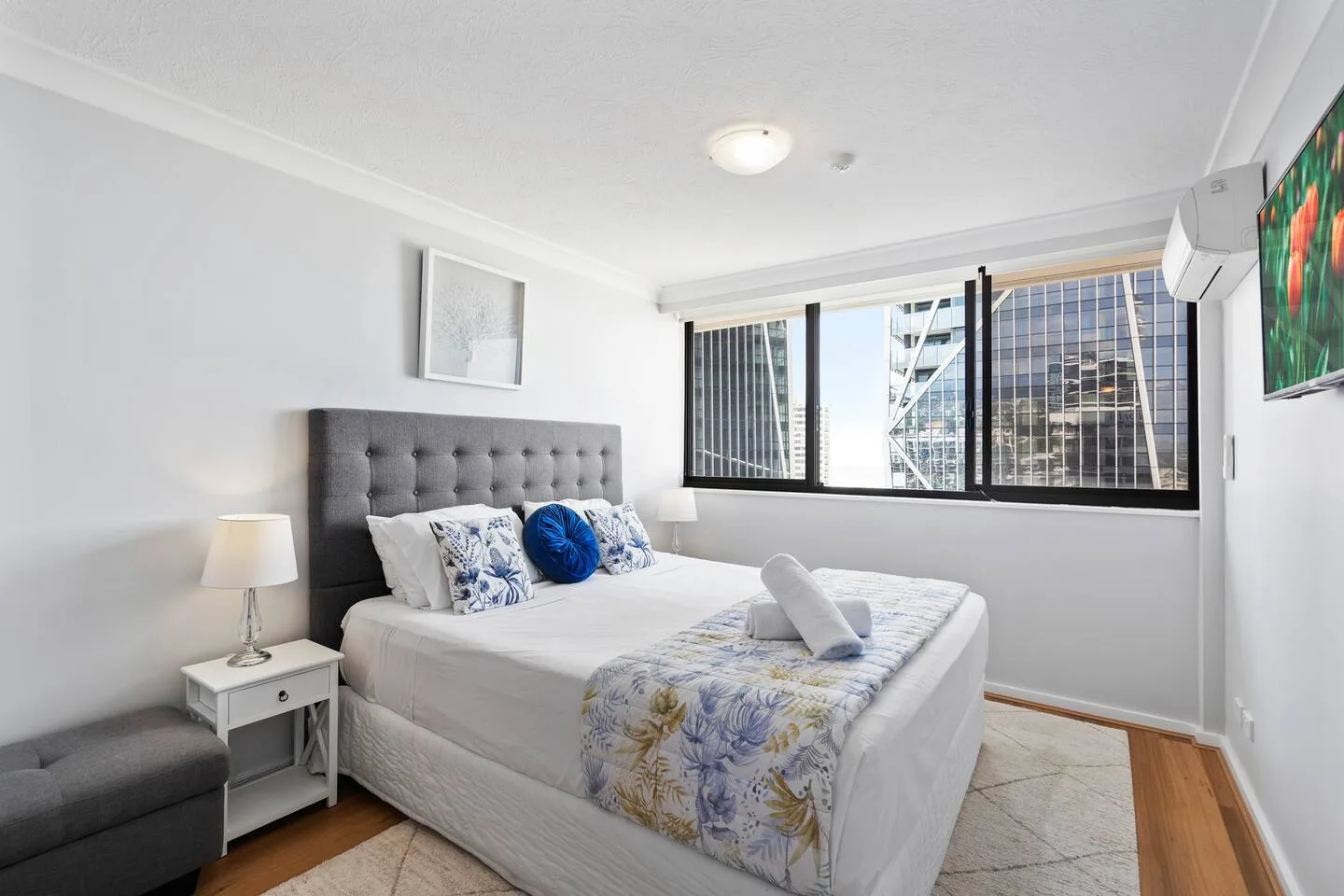 A bedroom with a made bed, bedside tables, and large windows overlooking nearby buildings.