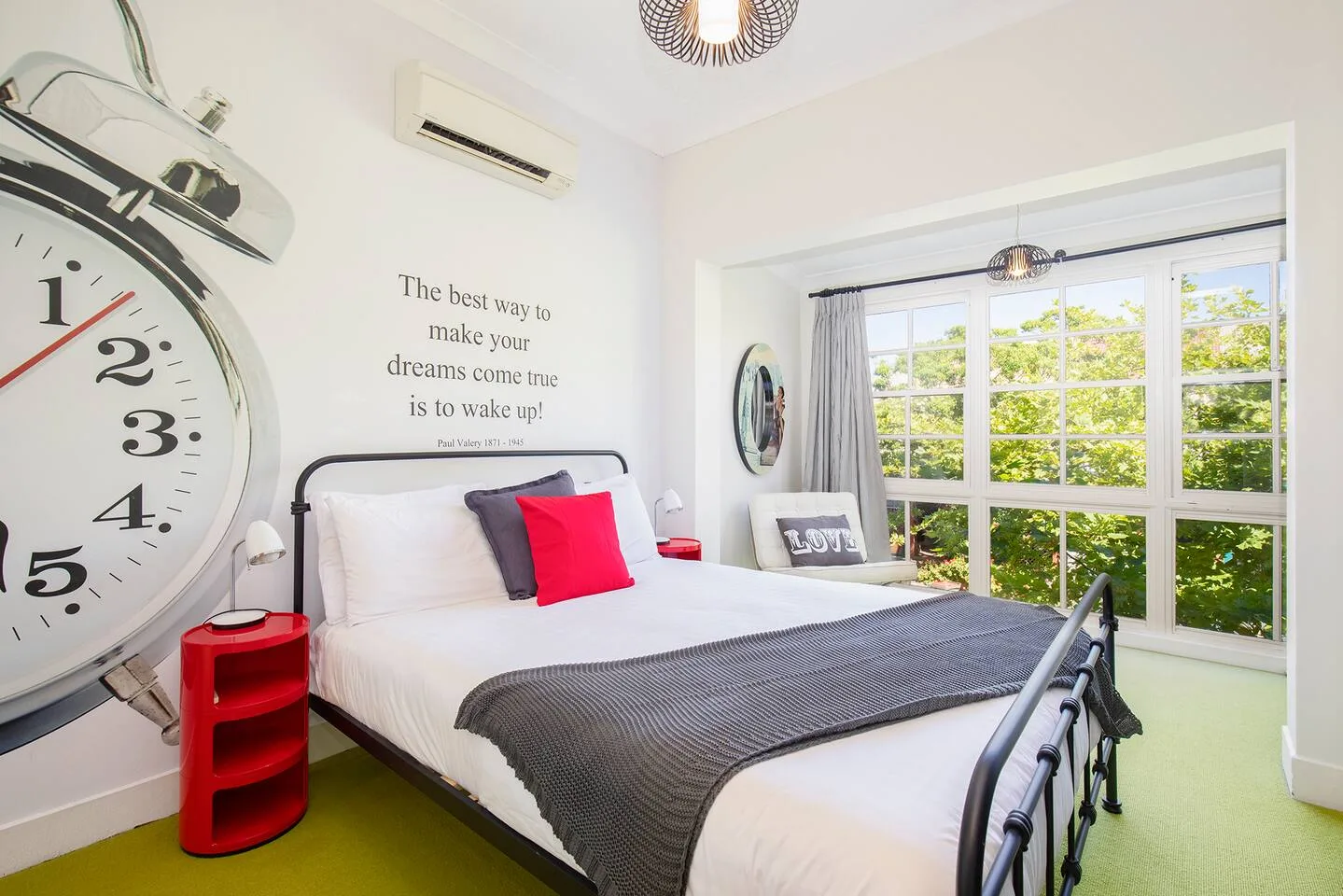 A bedroom with a bed beside large windows overlooking leafy greenery.