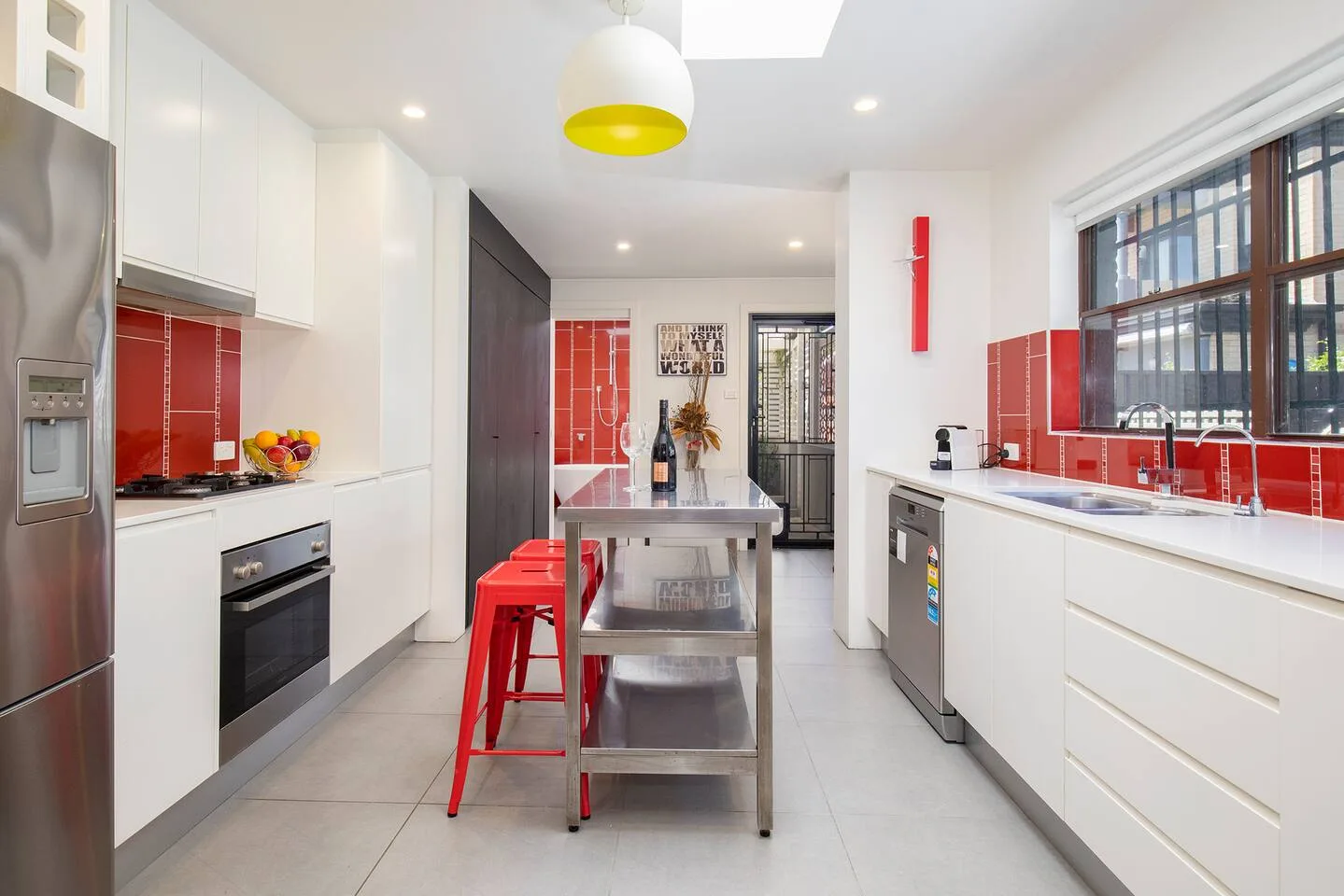 A bright modern kitchen has white cabinets, red tile accents, and a stainless-steel island.