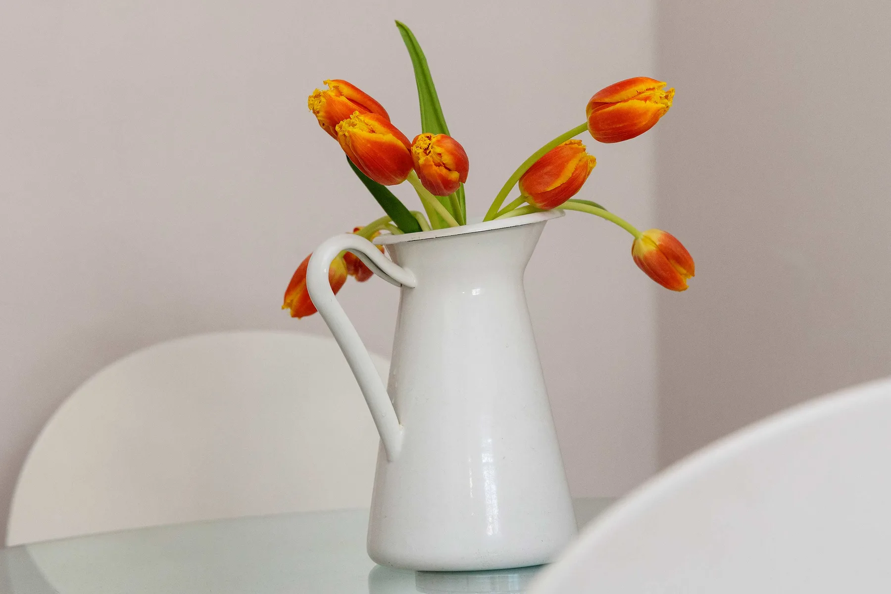 Una jarra blanca con flores naranjas está sobre una mesa de vidrio.
