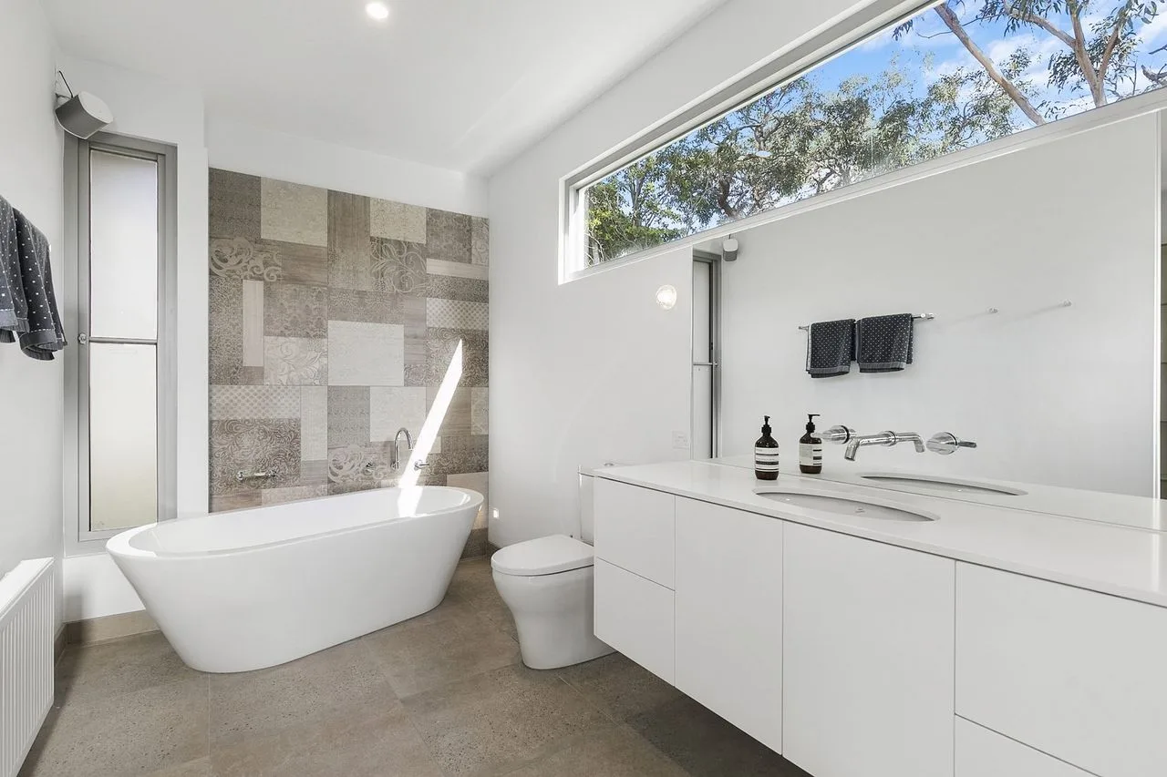 Un baño moderno y luminoso con bañera exenta y un largo tocador blanco.