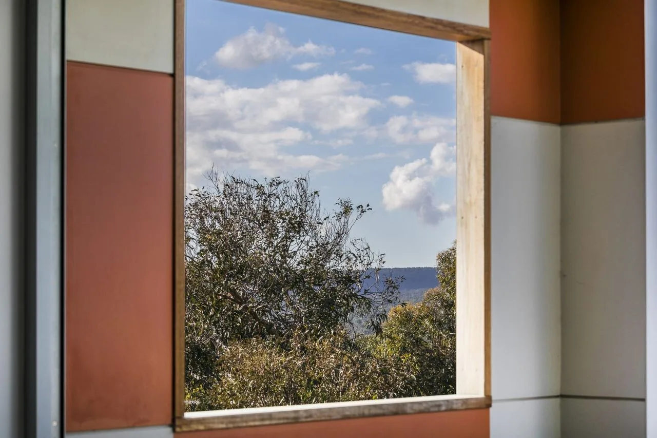 Una gran ventana enmarca árboles frondosos, colinas lejanas y un cielo nublado.