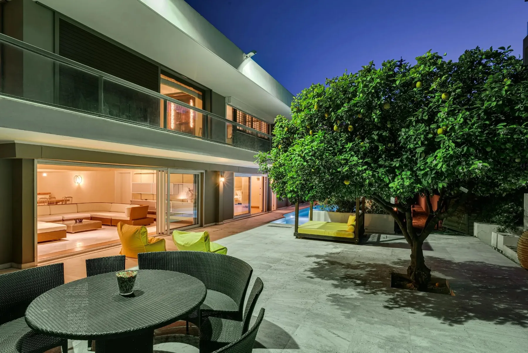 A modern house opens onto a paved courtyard with outdoor seating and a tree.