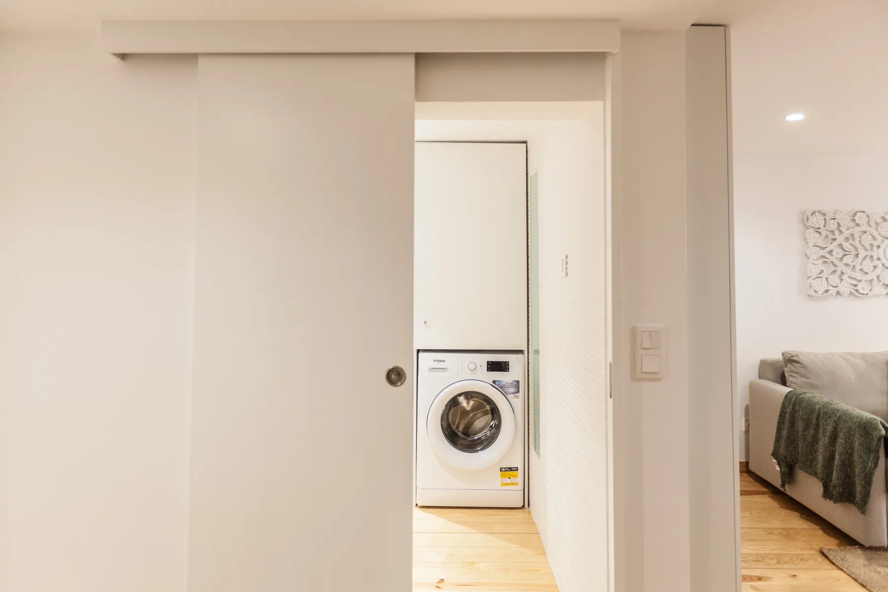 A laundry area with a front-loading washing machine beside a living room.