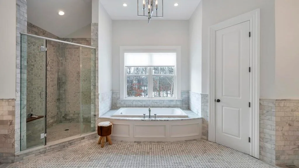 Un baño luminoso con una bañera bajo una ventana y una ducha de vidrio.