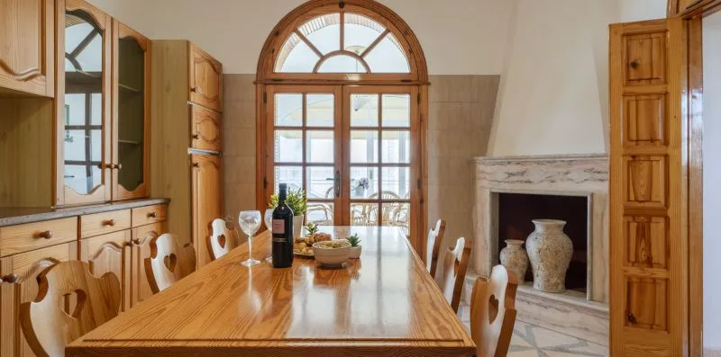 A dining room with a long wooden table, kitchen cabinets, and an arched window.