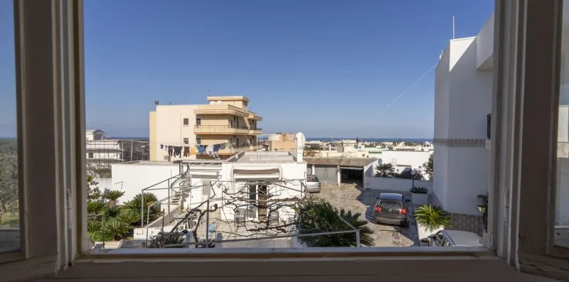 Vista desde una ventana hacia edificios cercanos, una zona de estacionamiento y el mar.
