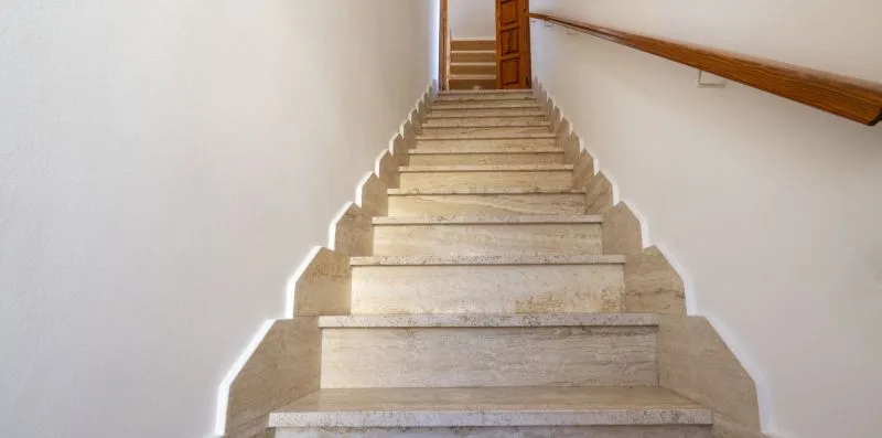 Una escalera de piedra sube hacia una puerta de madera, con un pasamanos junto a la pared.