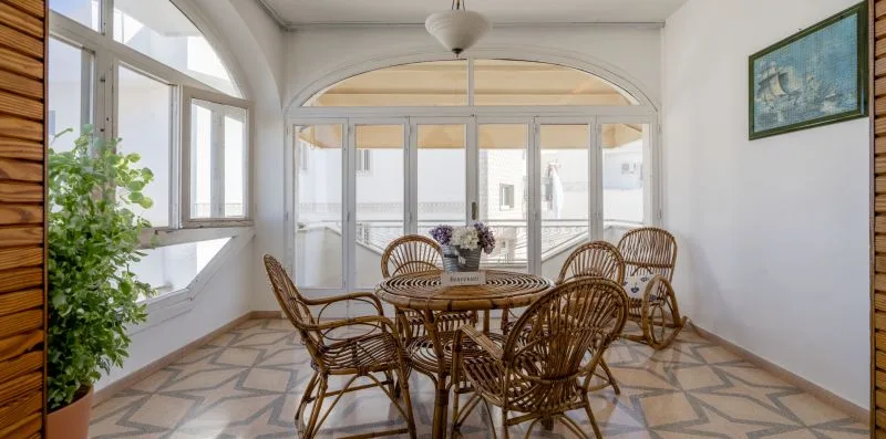 A bright sunroom with wicker seating around a round table and large windows.