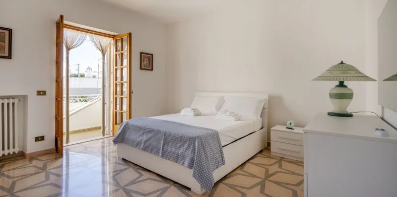 A bedroom with a neatly made bed beside open doors leading to a balcony.