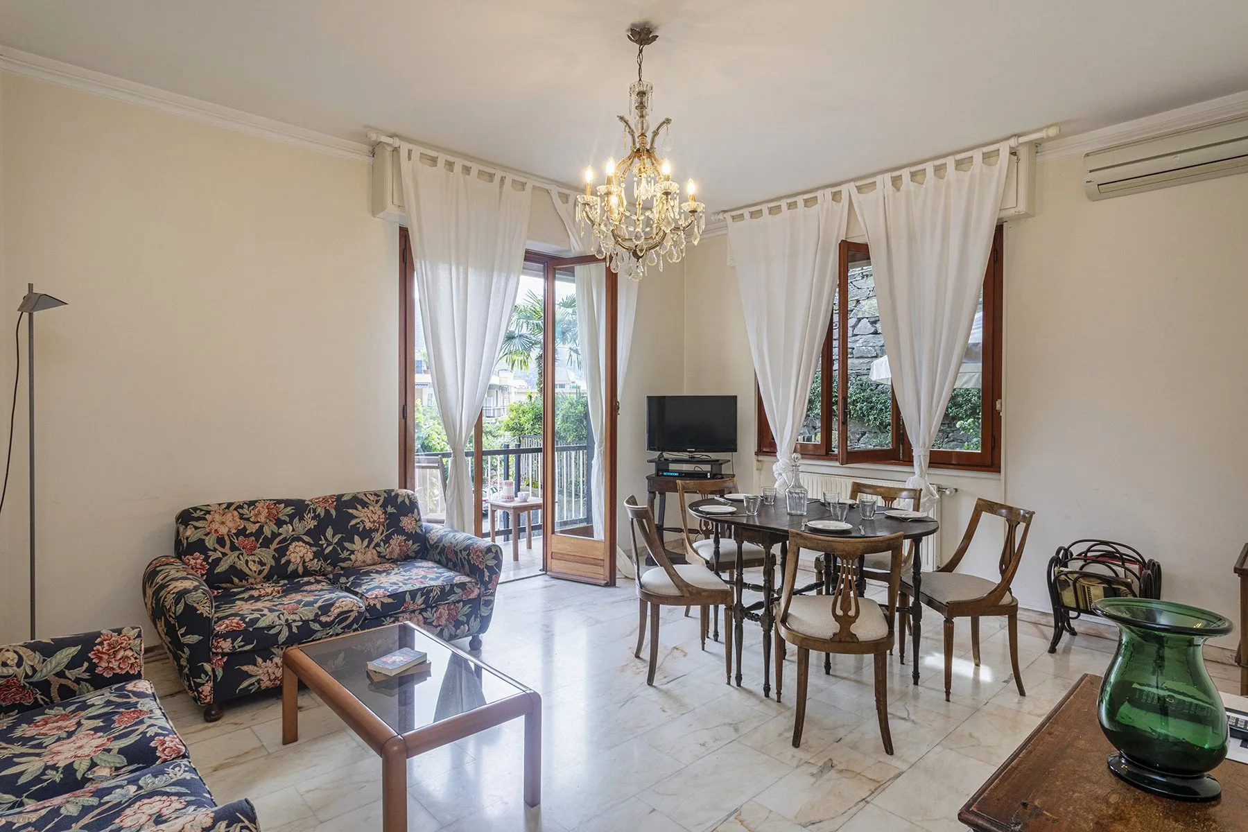Una sala de estar y comedor con asientos estampados, una mesa y ventanas altas.
