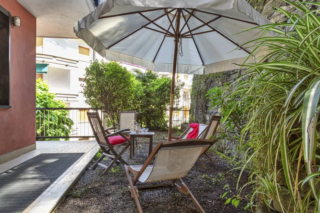Un patio al aire libre con asientos bajo una sombrilla grande, rodeado de plantas.