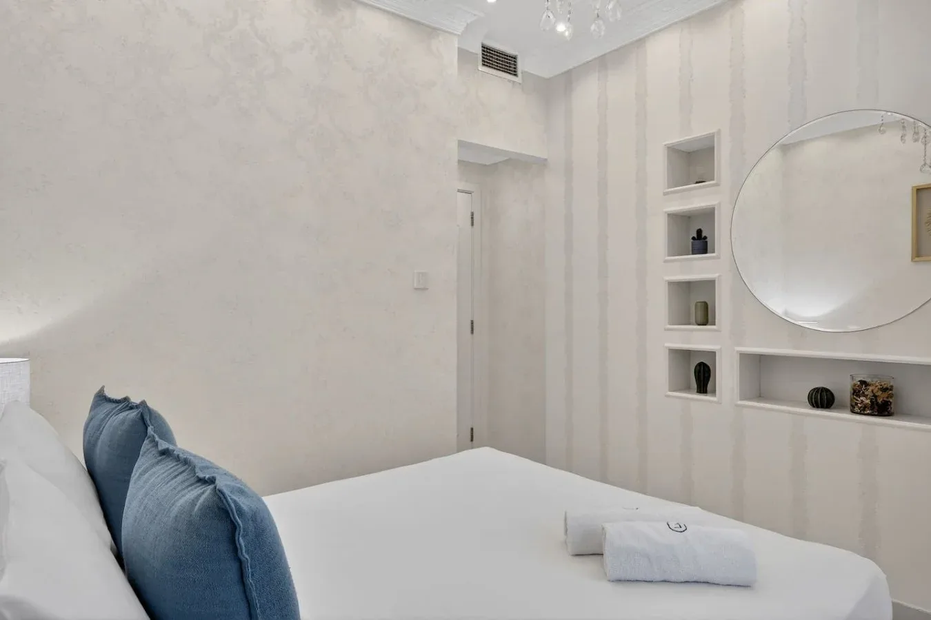 Un dormitorio con una cama blanca y almohadas azules junto a un espejo redondo.