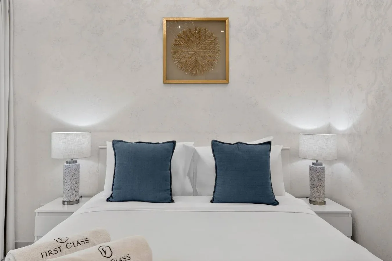 Un dormitorio ordenado muestra una cama blanca con almohadas azul oscuro y lámparas a los lados.