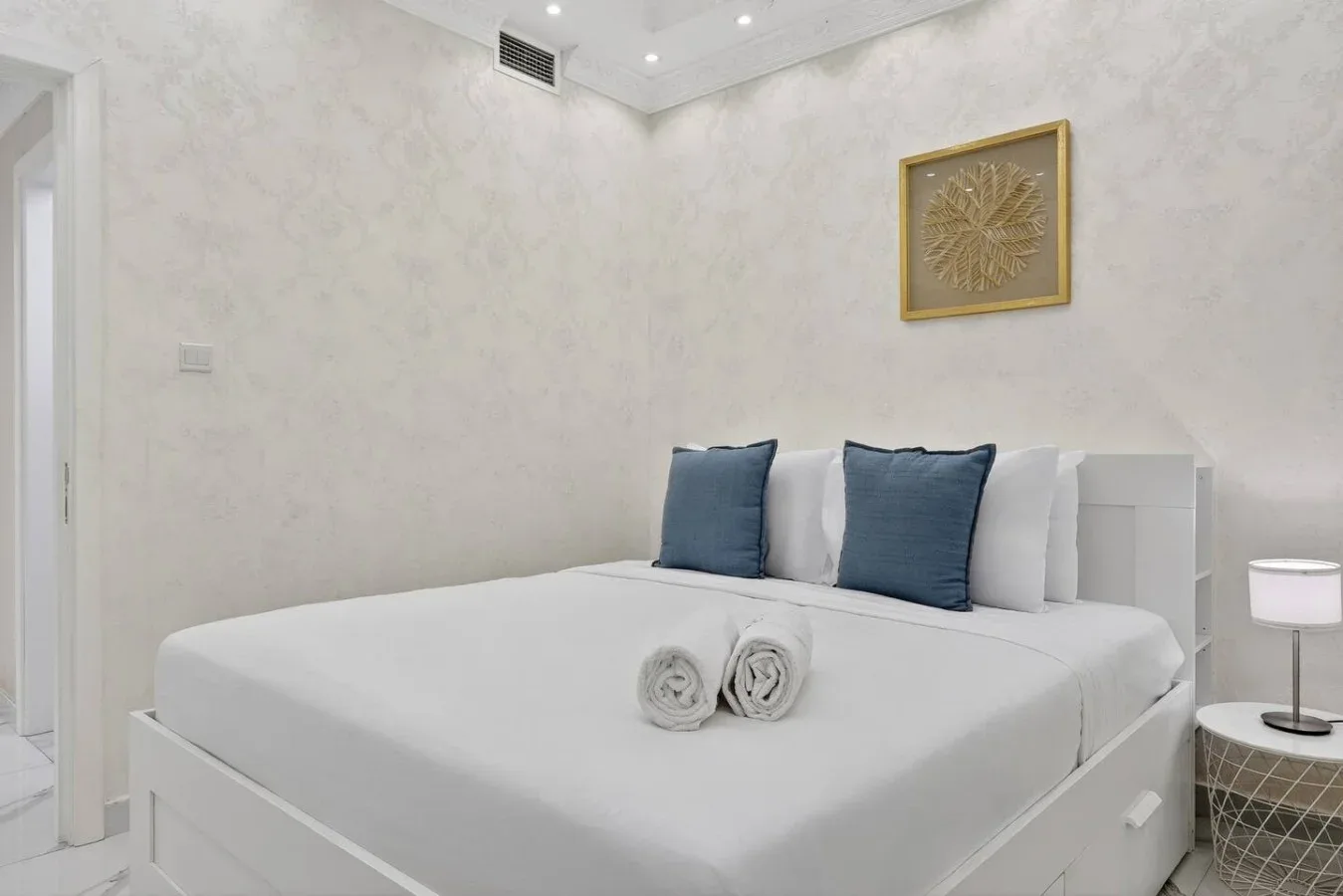 Un dormitorio luminoso con ropa de cama blanca, almohadas azules y dos toallas enrolladas.