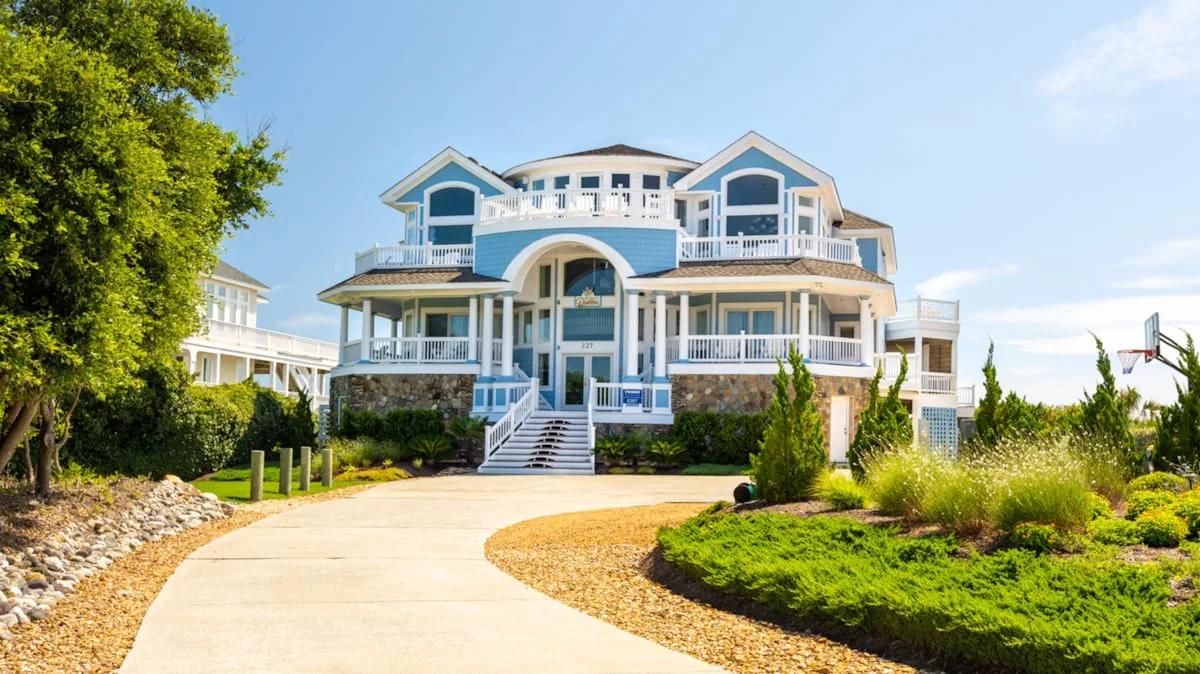 A large blue-and-white house stands beyond a curved driveway and landscaped garden.