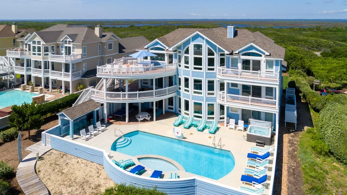 A large coastal house with balconies overlooks a swimming pool and surrounding greenery.