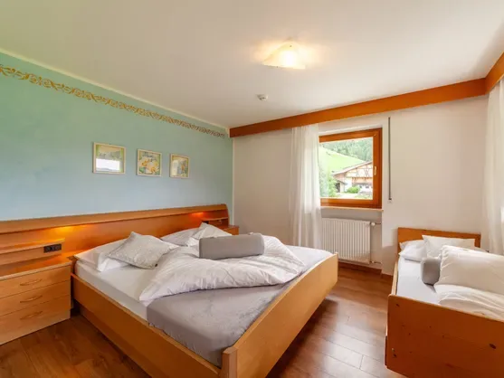 Habitación doble con cama de madera, cama supletoria y vistas desde la ventana a las verdes laderas de las montañas del valle de Sarentino