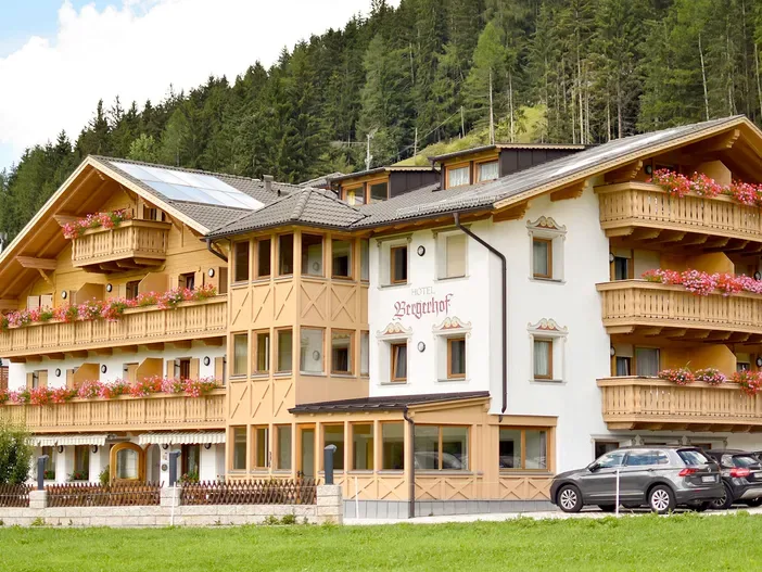Almhotel Bergerhof con balcones de madera y decoración floral, rodeado de bosque de montaña, en un día soleado