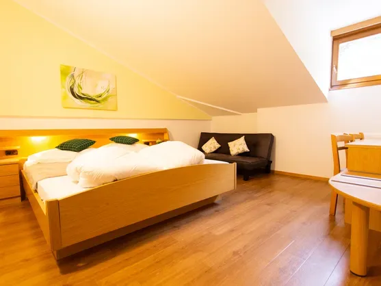 Habitación doble con cama de madera, pared amarilla, sofá y zona de estar bajo el techo inclinado