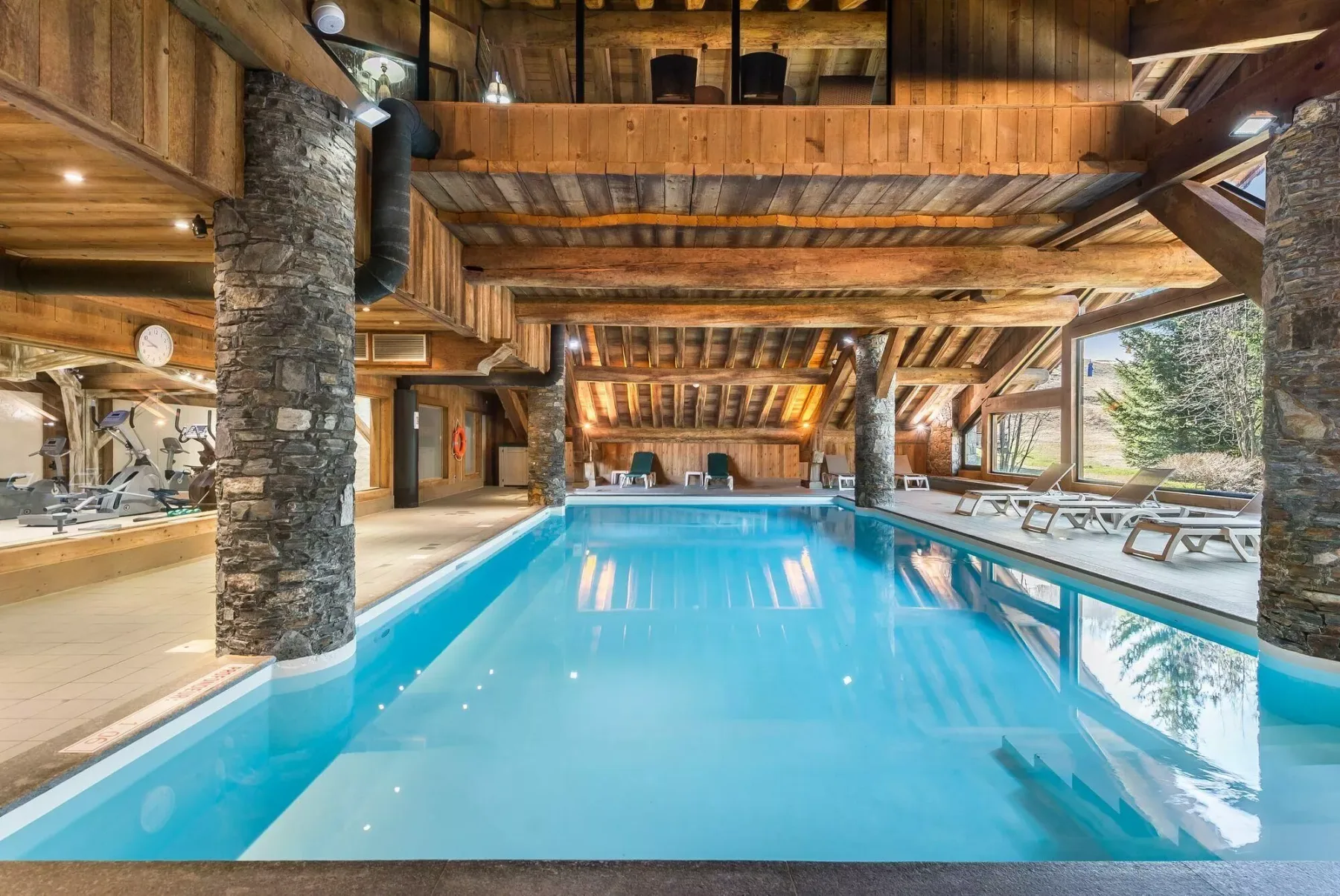 An indoor swimming pool beneath a timber ceiling, with lounge seating along the windows.