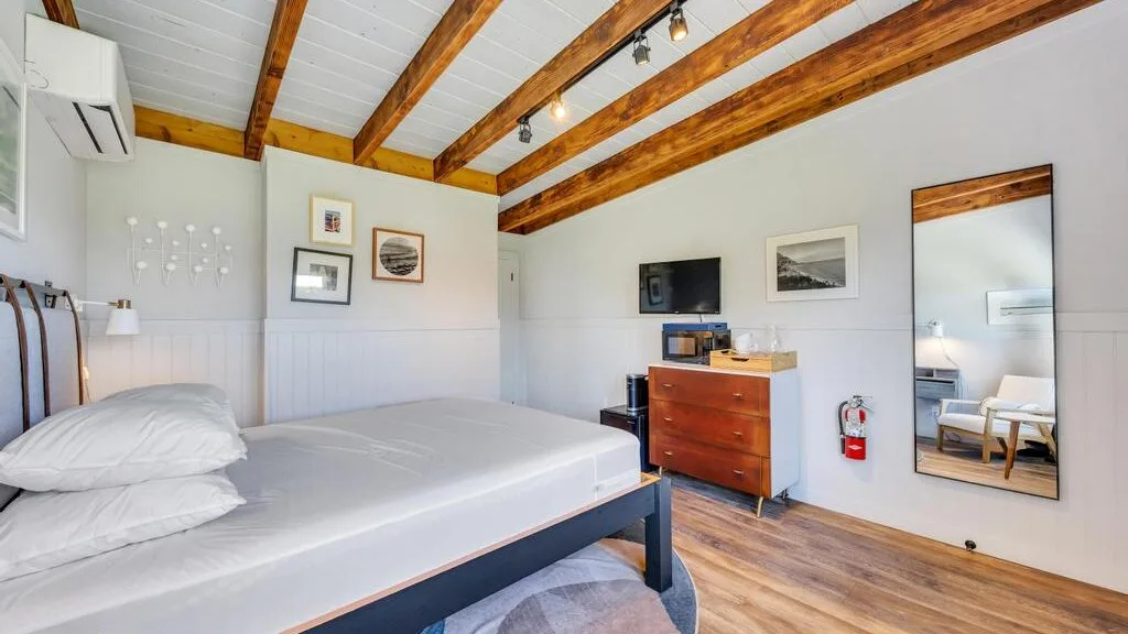 Un dormitorio luminoso tiene una cama, una cómoda de madera y vigas a la vista.