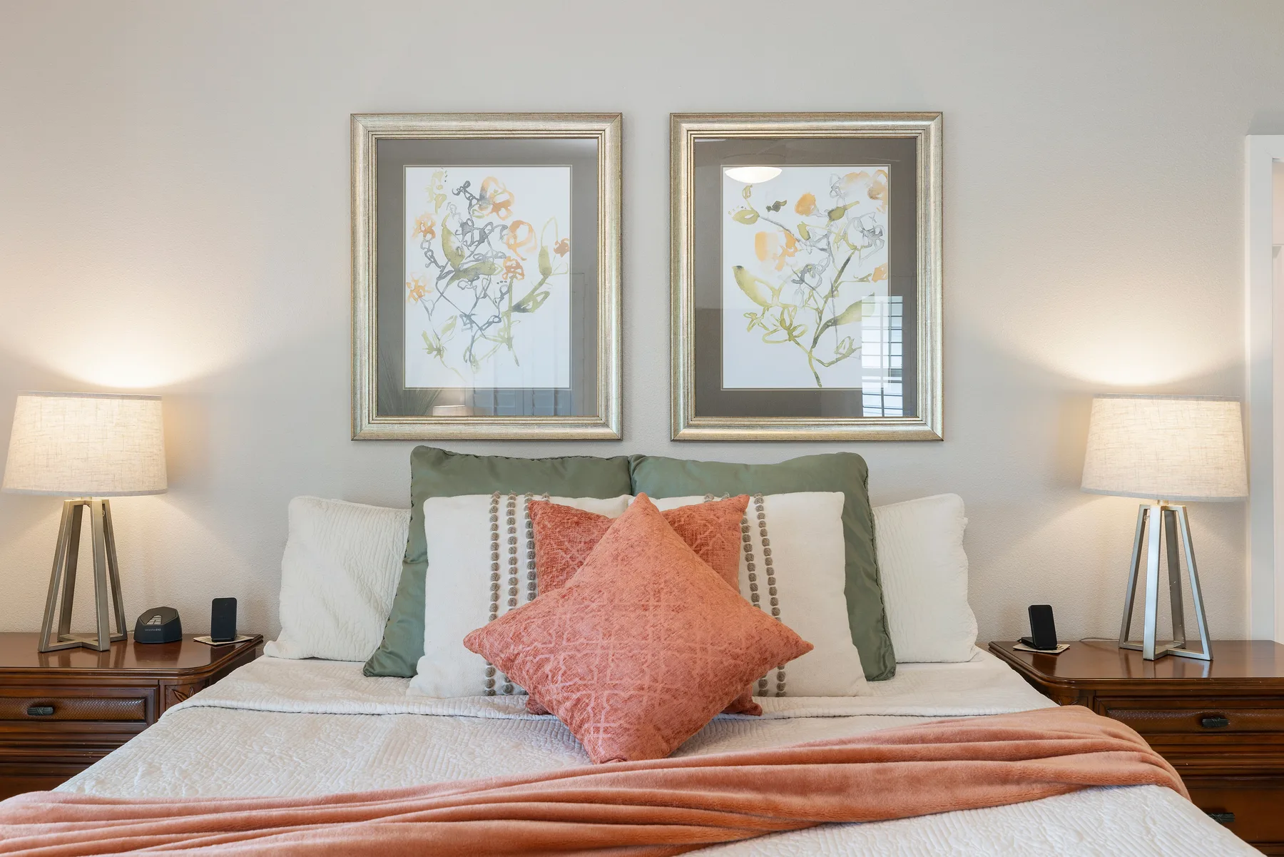 Una cama de dormitorio bien tendida, con cojines en capas y cuadros florales enmarcados encima.