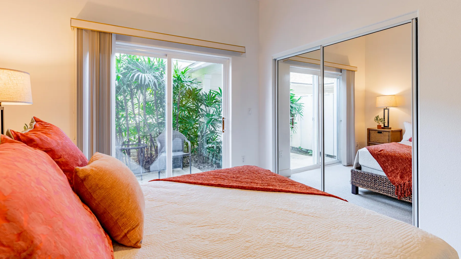Un dormitorio con una cama frente a puertas de armario espejadas y una puerta de vidrio hacia un patio frondoso.