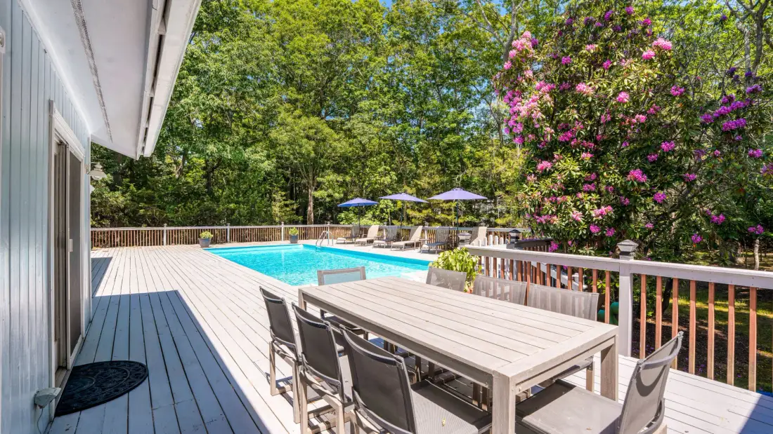 Una terraza exterior con mesa de comedor da a una piscina y árboles frondosos.