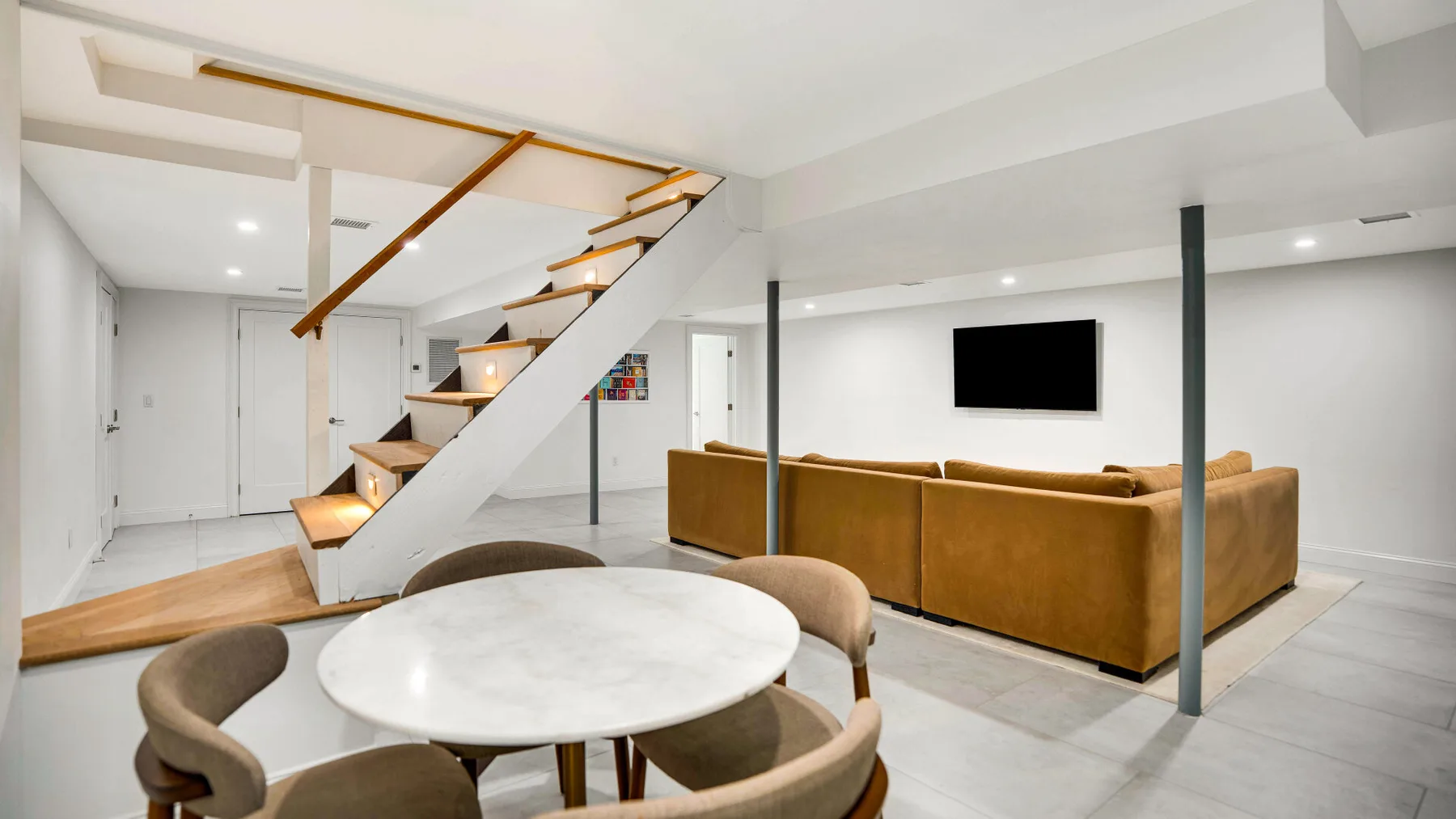 Una luminosa sala de estar con una escalera, asientos marrones y una mesa redonda.