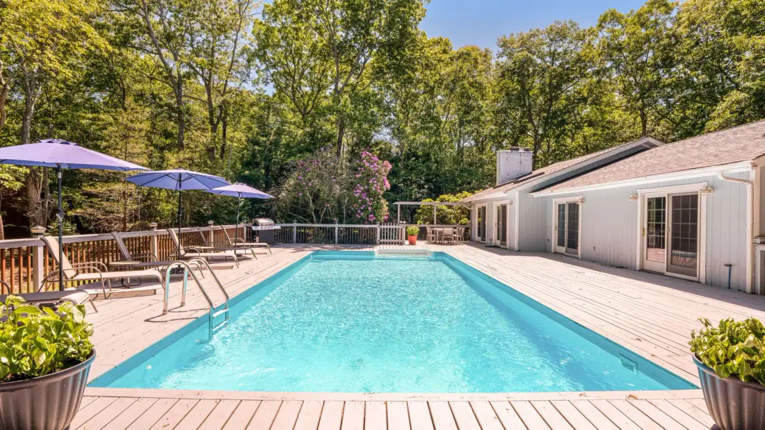 Una piscina al aire libre junto a un edificio blanco, con una terraza de madera y árboles.