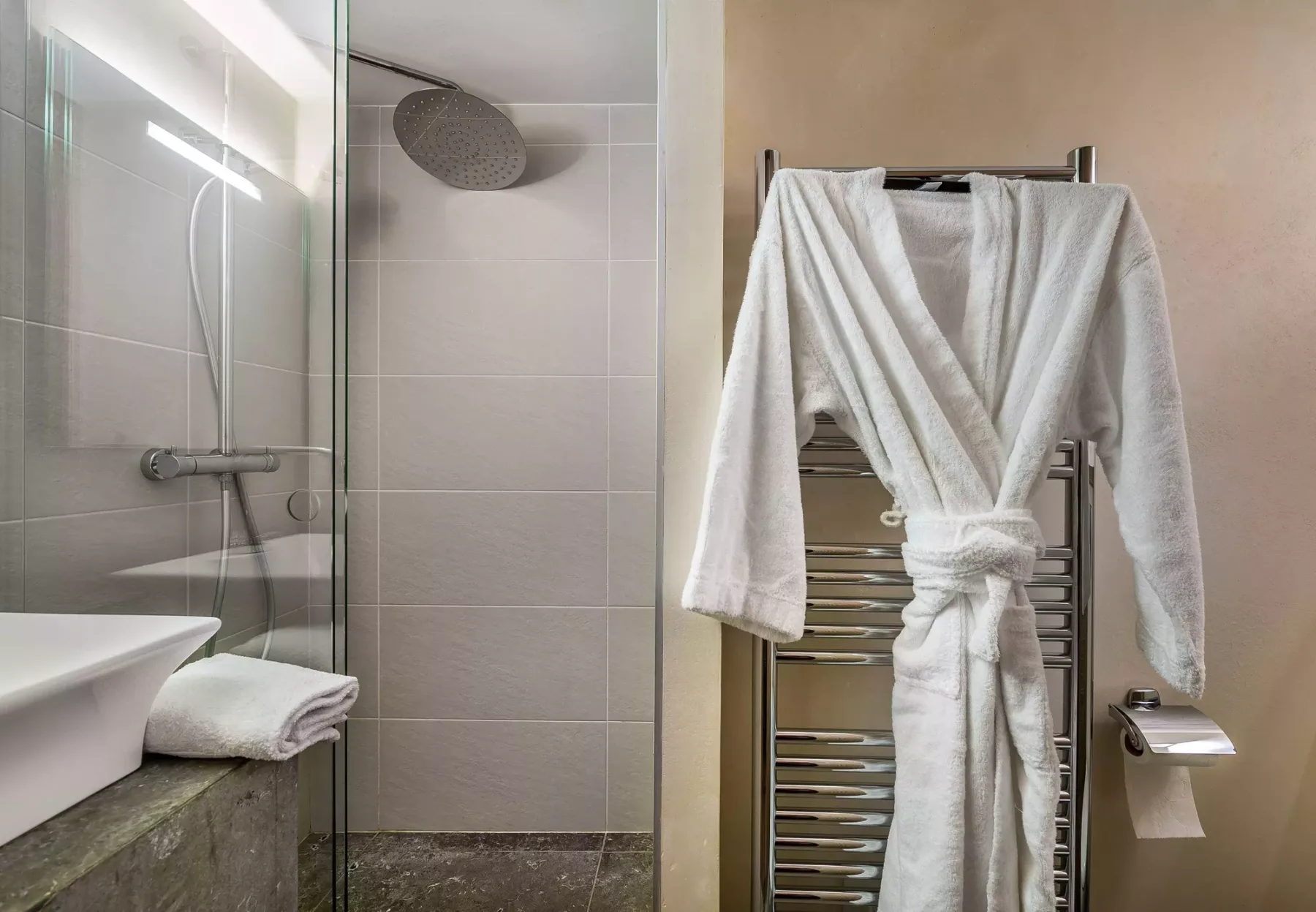 Un baño alicatado con una ducha de cristal y un albornoz blanco colgado en un toallero calefactable.