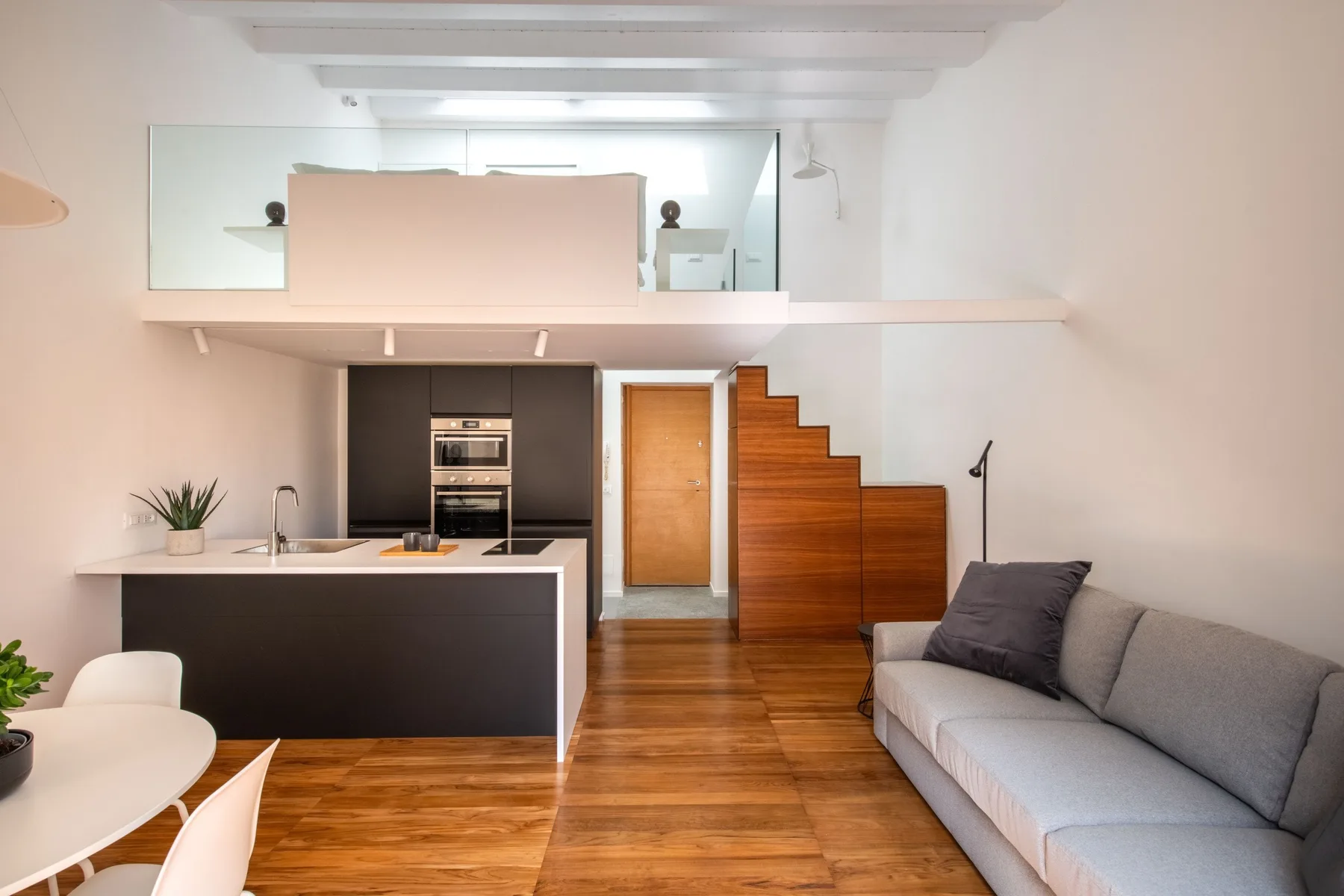A modern open-plan living area with a kitchen island, gray seating, and wooden stairs.