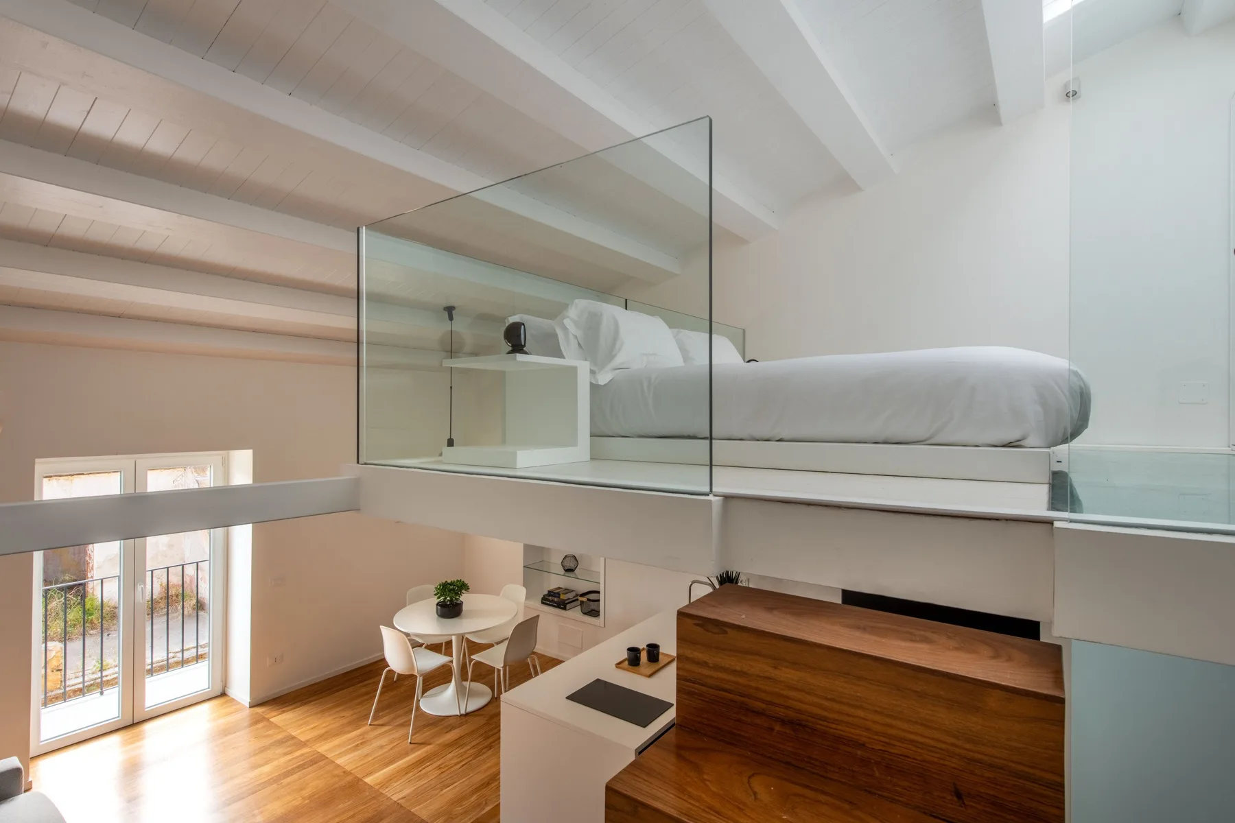 A bright loft bedroom overlooks a small dining area through a glass partition.