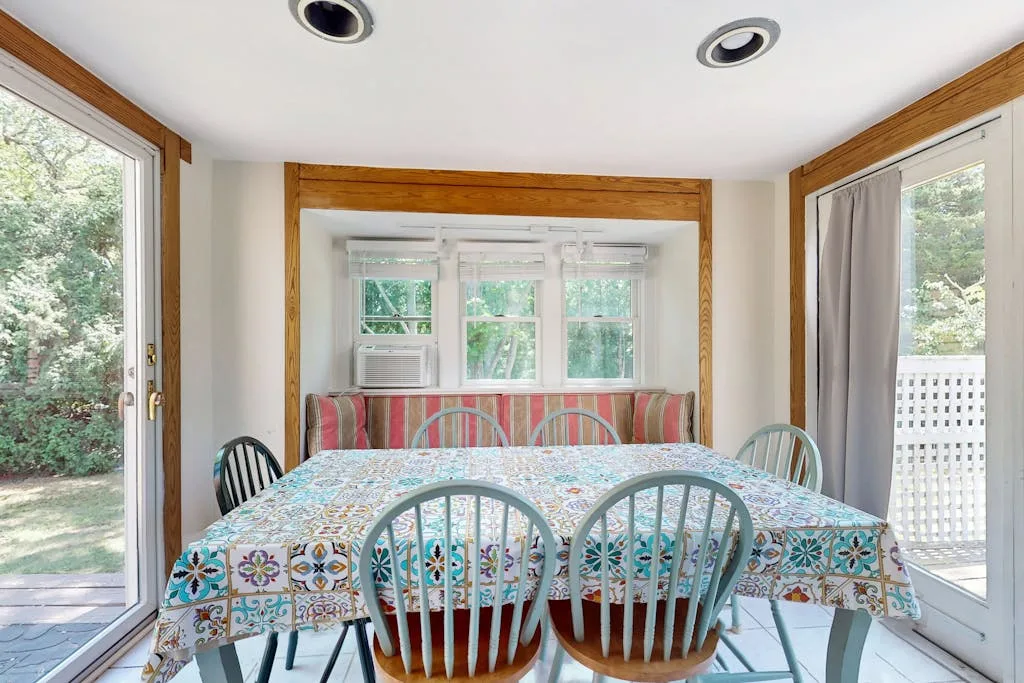 A sunroom dining area with a table covered by a patterned cloth and windows overlooking greenery.